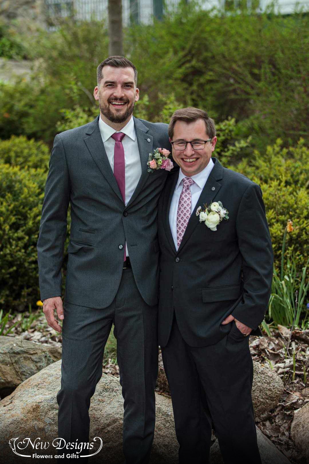 A groom and his groomsman stand side by side in a garden, sporting pocket square flowers instead of boutonnieres. the grooms in white and the groomsman's in a mix of whites and pinks
